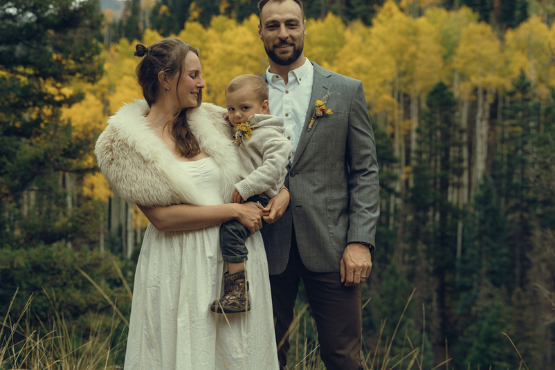 bride and groom pose with their son with yellow trees coloring the background