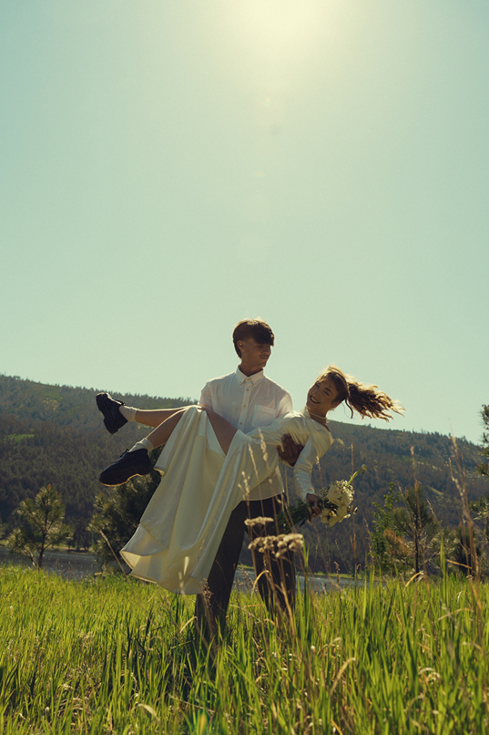 groom carries his bride in the field by the lake