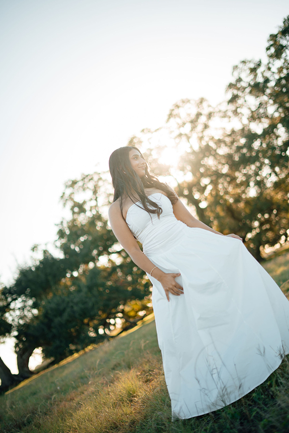 senior in white dress poses in a texas field as the suns gleems through the trees behind her