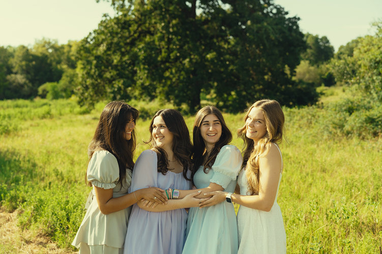 group of four girls laugh and embrace in a green field with sun beaming down on them