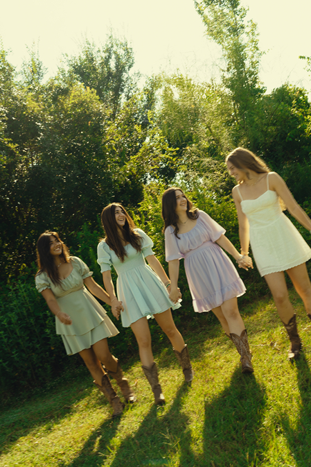 group of girls walk through a Texas field together holding hands and laughing during their senior session captured by Sasha Takes Pictures a Texas senior photographer 